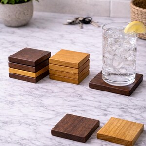 May include: Wooden square coasters in various shades of brown and tan. Some coasters are stacked, while others are placed individually. A glass of water with ice and a lemon slice sits on one coaster. The coasters are on a marble surface.