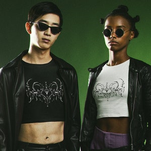 Puede incluir: Dos personas con chaquetas de cuero negro y tops cortos con un diseño gráfico plateado. La persona de la izquierda lleva pantalones de cuero negro y la persona de la derecha lleva pantalones morados.