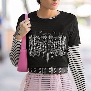 Può includere: Una maglietta nera con una grafica bianca di un design di fiamma stilizzato. La modella indossa una gonna di tulle rosa con strisce nere, una cintura nera con fibbie argentate e un berretto rosso con spille.