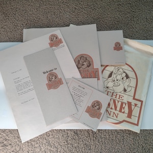 May include: A collection of vintage Disney Inn ephemera, including stationery, welcome cards, and a drawstring bag. The items feature the Disney Inn logo and illustrations of characters. The color palette includes beige, red, and brown.