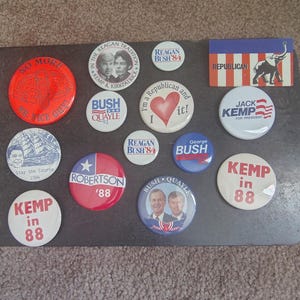 May include: A collection of vintage political campaign buttons and a small rectangular sign. The buttons feature images of candidates, slogans, and the Republican Party elephant logo. Colors include red, white, blue, and black.
