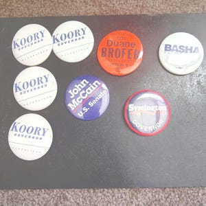 May include: A collection of political campaign buttons. The buttons feature names like "Koory Governor", "Duane Brofer", "Basha", "John McCain U.S. Senate", and "Symington Governor". The buttons are white, red, and blue.