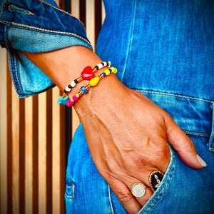 Puede incluir: Dos pulseras de cuentas coloridas con un dije de corazón y un dije de estrella. Las pulseras se usan en la muñeca de una persona. La persona lleva un mono de mezclilla azul y un anillo en el dedo.