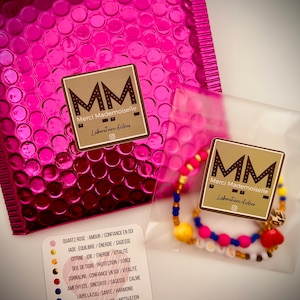 Puede incluir: Un primer plano de una pulsera de cuentas de colores con un dije en forma de coraz&oacute;n. La pulsera est&aacute; empaquetada en una bolsa de pl&aacute;stico transparente con un sobre de burbujas rosa en el fondo. El empaque incluye una tarjeta con el texto "MM Merci Mademoiselle Laboratoire d'id&eacute;es" y una lista de piedras preciosas y sus significados asociados.