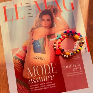Puede incluir: Una portada de revista con el t&iacute;tulo "LE MAG" y el texto "MODE assum&eacute;e" en letras grandes y en negrita. La portada presenta a una mujer con un top de crochet de colores y un bolso de paja con el texto "ISABEL MARANT". La revista est&aacute; sobre una superficie de madera con una pulsera de cuentas de colores con abalorios.