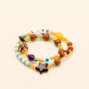 Peut inclure: Un bracelet multi-rangs avec des perles en bois, des perles vertes et dorées, un charme en forme d'étoile, un charme œil maléfique et une perle à imprimé léopard. Le bracelet épelle "LOVE" avec des perles de lettres blanches.