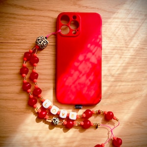 Puede incluir: Una funda de teléfono roja con una correa de teléfono con cuentas. La correa tiene cuentas rojas y naranjas, un dije de estampado de leopardo y cuentas de letras blancas que deletrean "LOVE".