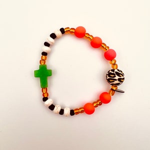 Puede incluir: Un brazalete de cuentas de colores con un dije de cruz verde, cuentas de color naranja y blanco, y una cuenta de estampado de leopardo en blanco y negro.