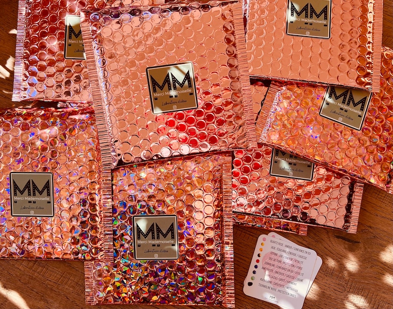 Puede incluir: Una pila de sobres de burbujas de color oro rosa con el logotipo "MM Merci Mademoiselle" impreso en cada uno. Una peque&ntilde;a tarjeta con texto en franc&eacute;s es visible en la esquina inferior derecha.