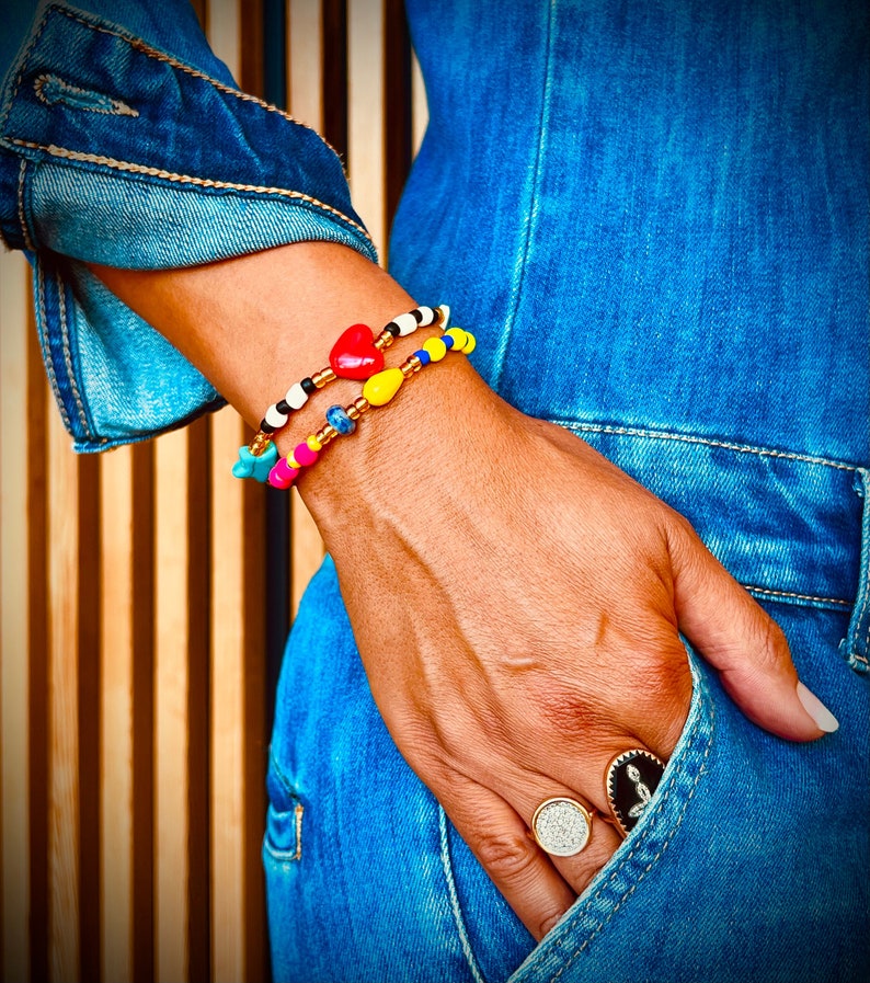 Puede incluir: Dos pulseras de cuentas coloridas con un dije de coraz&oacute;n y un dije de estrella. Las pulseras se usan en la mu&ntilde;eca de una persona. La persona lleva un mono de mezclilla azul y un anillo en el dedo.