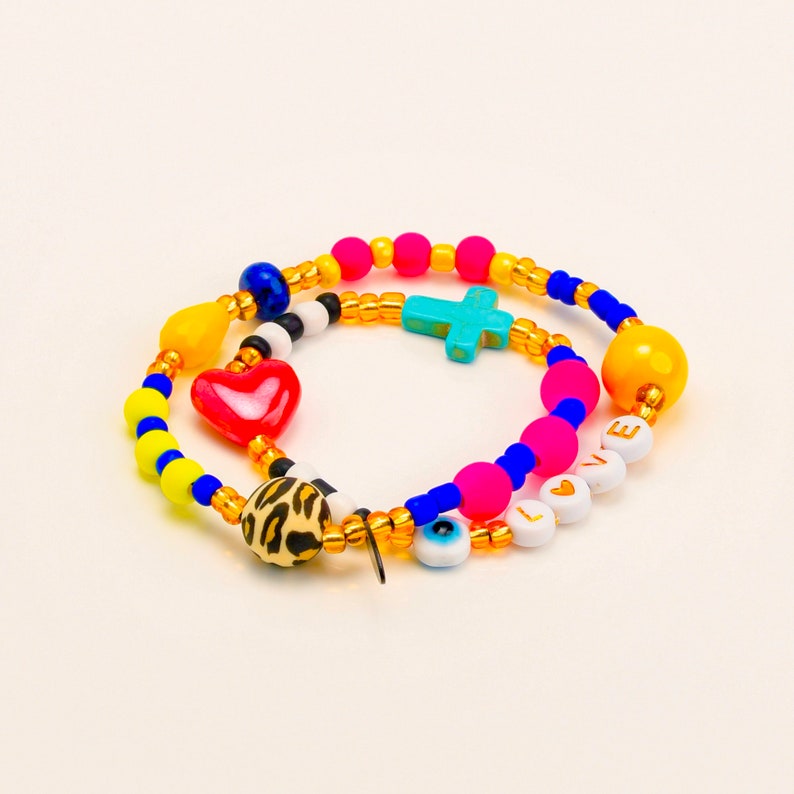 Puede incluir: Un brazalete de cuentas de colores con un dije de coraz&oacute;n, cruz y ojo. El brazalete tambi&eacute;n tiene la palabra "LOVE" escrita en letras doradas sobre cuentas blancas.