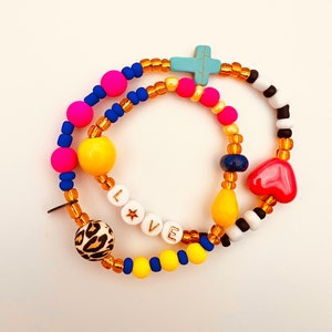 Puede incluir: Un brazalete de cuentas de colores con un dije de cruz, un dije de coraz&oacute;n y la palabra "LOVE" escrita en cuentas blancas con letras doradas. El brazalete est&aacute; hecho con cuentas amarillas, rosas, azules y marrones.