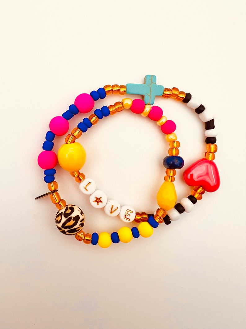 Puede incluir: Un brazalete de cuentas de colores con un dije de cruz, un dije de coraz&oacute;n y la palabra "LOVE" escrita en cuentas blancas con letras doradas. El brazalete est&aacute; hecho con cuentas amarillas, rosas, azules y marrones.
