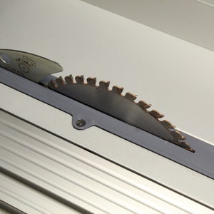 Może przedstawiać: Ostrze piły tarczowej z ząbkowaną krawędzią jest częściowo widoczne w szarej obudowie. Ostrze jest srebrne z szeregiem zębów wzdłuż krawędzi. Obudowa jest zamontowana na jasnoszarej powierzchni.