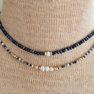 May include: A double-strand necklace featuring one layer of faceted dark blue beads with a central white pearl and gold spacers, and a second layer of multi-colored small beads accented by three central white pearls and gold-toned metal beads.