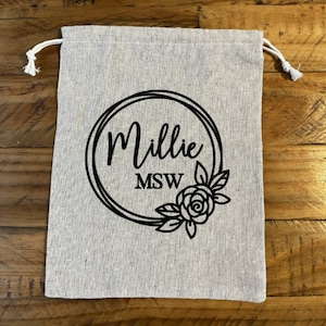 May include: A white canvas drawstring bag with a black circle design. The circle contains the text "Millie MSW" and a black floral design.