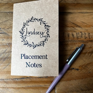 May include: A tan paper notebook featuring a black printed wreath design, the name Lindsey, and the text Placement Notes. A metallic mauve retractable ballpoint pen with a silver clip and tip is positioned diagonally next to the notebook.