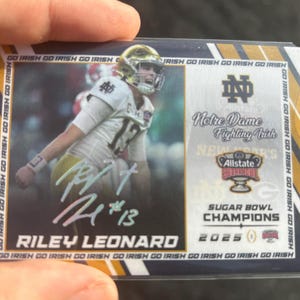 May include: A blue and gold Notre Dame Fighting Irish football card featuring Riley Leonard, a player in the 2025 Sugar Bowl Champions. The card has a gold and black border with the words "Go Irish" repeated around the edge.