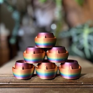 Op de afbeelding: Een stapel van zes regenboogkleurige keramische potten, elk met een iets andere tint van iriserende afwerking. De potten zijn in een piramidevorm gerangschikt, met de grootste pot onderaan en de kleinste pot bovenaan.
