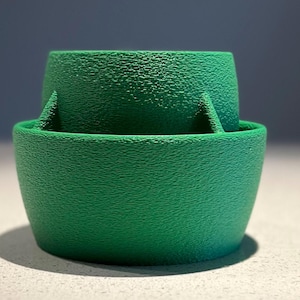 Puede incluir: Una maceta de plástico verde con una superficie texturizada. La maceta tiene un borde elevado alrededor de la parte superior y un borde más pequeño alrededor de la parte inferior.