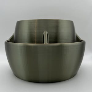 Op de afbeelding: Een tweedelige, olijfgroene, 3D-geprinte plantenbak. Het buitenste deel is een brede, komvormige basis. Het binnenste deel is een kleinere, cilindrische container met een verticale handgreep. De plantenbak heeft een getextureerde, metallic uitstraling.