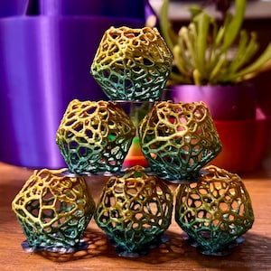 Pode incluir: Seis pequenas esculturas geométricas vazadas, com gradiente de amarelo, verde e azul. Estão dispostas sobre uma superfície de madeira, cada uma sobre uma base circular transparente. O fundo apresenta vasos roxos e rosas e vegetação.