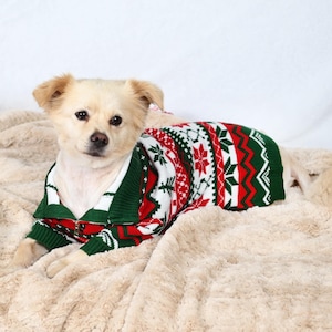 May include: A small, light-colored dog wearing a Christmas-themed sweater. The sweater features a pattern of red, green, and white snowflakes and geometric designs. The dog is lying on a soft, beige blanket.