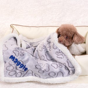 May include: A gray and white dog bed blanket with a paw print design. The blanket has a white border and the name "Maggie" embroidered in blue on the blanket. A small brown dog is curled up on the blanket.