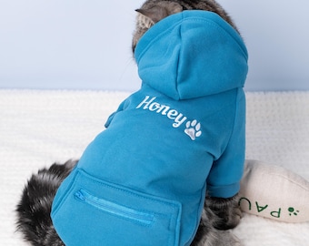 May include: A blue pet hoodie with a hood and a front pocket. The word "Honey" is embroidered on the back in white, along with a paw print. The hoodie is on a gray tabby cat.