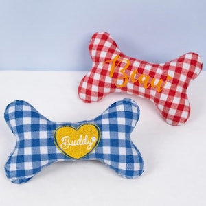Op de afbeelding: Twee hondenbotvormige pluche speeltjes. E&eacute;n is rood en wit geblokt met de naam "Beau" in oranje geborduurd. De andere is blauw en wit geblokt met een geel hart met "Buddy".