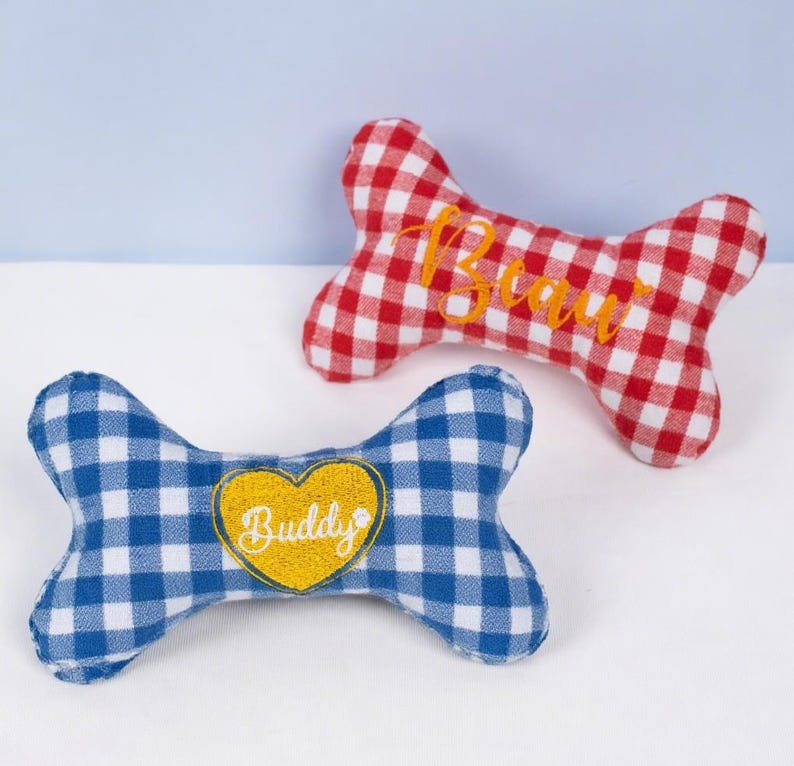 Op de afbeelding: Twee hondenbotvormige pluche speeltjes. E&eacute;n is rood en wit geblokt met de naam "Beau" in oranje geborduurd. De andere is blauw en wit geblokt met een geel hart met "Buddy".