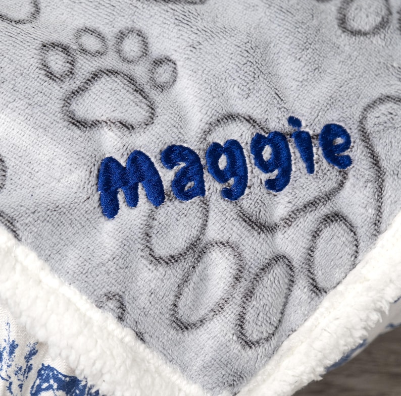 May include: Gray and white pet bed with a blue embroidered paw print pattern and the name "Maggie" in blue.