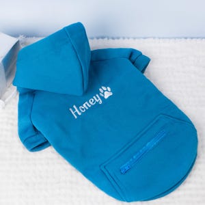 May include: A turquoise dog hoodie with a hood and a zippered pocket. The word "Honey" is embroidered in white, along with a paw print. The hoodie is made of a soft material.