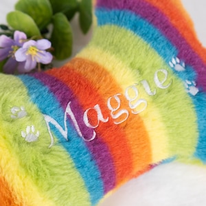 Op de afbeelding: Een regenboogkleurig pluche hondenbot met de naam "Maggie" in het wit geborduurd. Het bot heeft een levendig kleurenspectrum en pootafdrukdetails, perfect voor een huisdier.