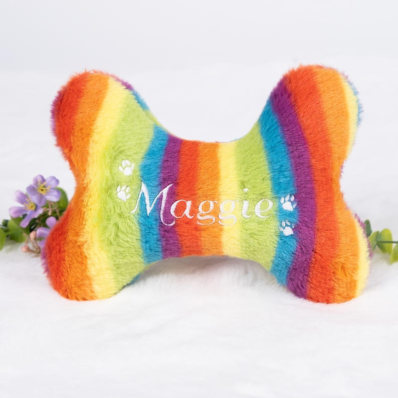 Op de afbeelding: Een regenboog gestreept hondenbotvormig pluche speelgoed. Het speelgoed heeft de naam "Maggie" geborduurd in wit script, samen met pootafdrukdetails. Het pluche is een levendige mix van kleuren, waaronder rood, oranje, geel, groen, blauw en paars.