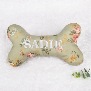 Op de afbeelding: Een hondenbotvormig kussen van saliegroene stof, geborduurd met de naam "SADIE" in wit. Het kussen is versierd met een bloemenpatroon in roze, wit en geel.