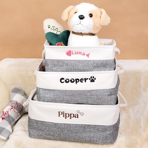 May include: Three gray and white fabric storage bins stacked, each with a name: Luna, Cooper, and Pippa. A plush dog toy and a green heart-shaped toy are in the top bin. A stuffed dog toy is also in the top bin. A bone-shaped toy is on the left.