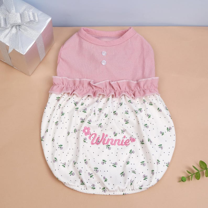 Peut inclure: Un v&ecirc;tement pour animal de compagnie avec un corsage rose en maille gaufr&eacute;e, deux boutons blancs et une taille &agrave; volants. La partie inf&eacute;rieure est blanche avec des pois noirs et des petites fleurs, portant le nom Winnie brod&eacute; en rose.