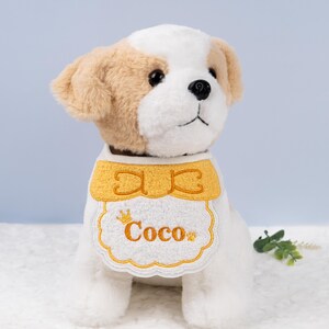 May include: White plush dog wearing a white and yellow embroidered bib with a crown and the name "Coco" on it.