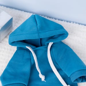 May include: A vibrant turquoise hoodie with a white drawstring and a matching hood. The hoodie is made of a soft material and is displayed on a white, fluffy surface. The sleeves have a ribbed cuff.