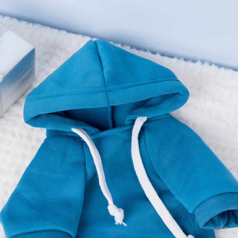 May include: A vibrant turquoise hoodie with a white drawstring and a matching hood. The hoodie is made of a soft material and is displayed on a white, fluffy surface. The sleeves have a ribbed cuff.