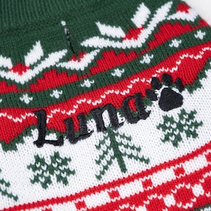 May include: Close-up of a green, red, and white knitted dog sweater. The sweater features a repeating pattern of snowflakes and trees. The name "Luna" is embroidered in black, along with a black paw print.