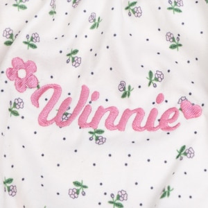 Peut inclure: Un tissu blanc orn&eacute; d'un motif r&eacute;p&eacute;titif de minuscules fleurs violettes et de petits points sombres. Le pr&eacute;nom Winnie est brod&eacute; en lettres cursives roses au centre, encadr&eacute; par une fleur rose brod&eacute;e &agrave; gauche et un c&oelig;ur rose &agrave; droite.