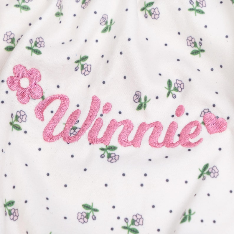 Peut inclure: Un tissu blanc orn&eacute; d'un motif r&eacute;p&eacute;titif de minuscules fleurs violettes et de petits points sombres. Le pr&eacute;nom Winnie est brod&eacute; en lettres cursives roses au centre, encadr&eacute; par une fleur rose brod&eacute;e &agrave; gauche et un c&oelig;ur rose &agrave; droite.