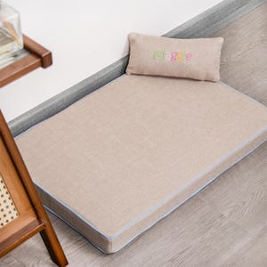 May include: A beige rectangular dog bed with a blue trim. The bed has a matching pillow with the name "Maggie" embroidered on it in colorful letters.