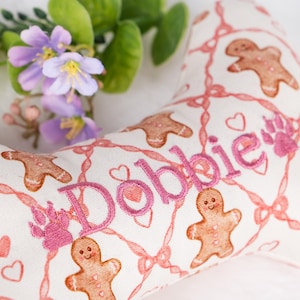 Op de afbeelding: Een botvormig hondenspeeltje met een witte achtergrond, met een patroon van roze peperkoekmannetjes, hartjes en linten. De naam "D&ouml;bbie" is roze geborduurd. Een pootafdruk is ook roze geborduurd.