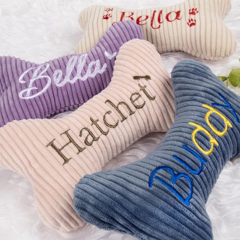 Op de afbeelding: Vier corduroy hondenbot speelgoed in tinten paars, beige en blauw. Elk bot heeft een geborduurde naam in een contrasterende kleur: "Bella", "Hatchet", "Buddy" en "Kella". De speeltjes zijn ontworpen voor huisdieren.