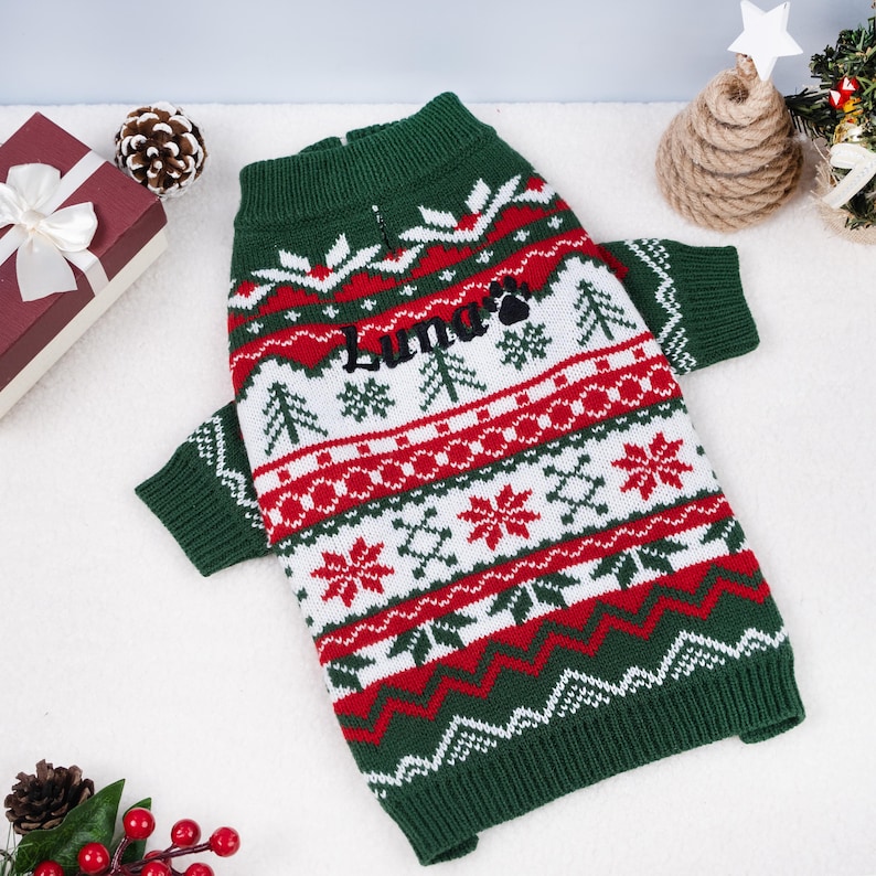 May include: A green, red, and white Christmas-themed dog sweater. The sweater features a pattern of snowflakes, trees, and zigzags. The name "Luna" is embroidered on the front. The sweater has a high collar and sleeves.