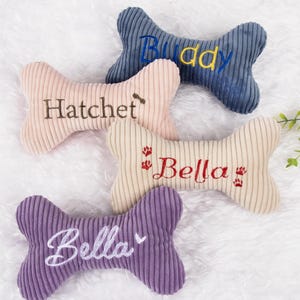 Op de afbeelding: Vier corduroy hondenbot speelgoed in blauw, roze, beige en paars. Elk bot heeft een geborduurde naam: "Buddy", "Hatchet" en "Bella". Deze pluche speelgoed zijn ontworpen voor honden.