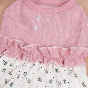 Peut inclure: Un v&ecirc;tement sans manches avec un corsage en maille gaufr&eacute;e rose poudr&eacute; dot&eacute; de deux petits boutons blancs &agrave; l'encolure. Un volant horizontal s&eacute;pare le corsage d'une jupe cr&egrave;me orn&eacute;e d'un motif de minuscules points noirs et de petites fleurs.